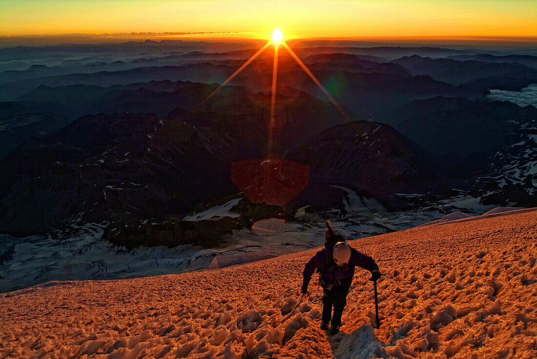 A climber ascends the Emmons-Winthrop Glacier route with an ice axe and a rope on Mount Rainier as the sun just peaks over the horizon just after sunrise
