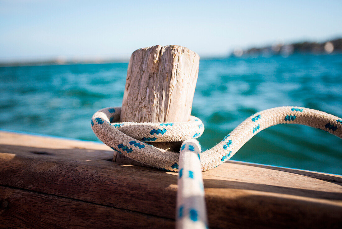 A rope wrapped around an old wood peg on a wooden sailboat adrift in blue water.