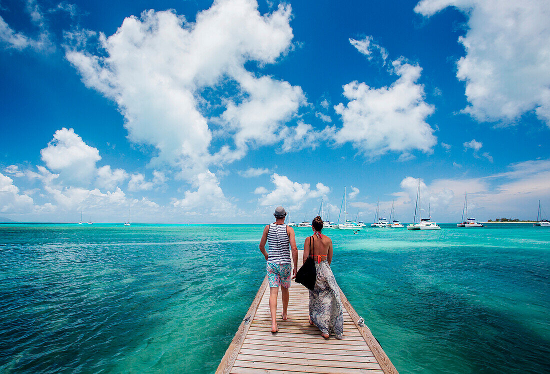 A couple walks down the long dock of Anegada Island surrounded by teal sea and blue sky.