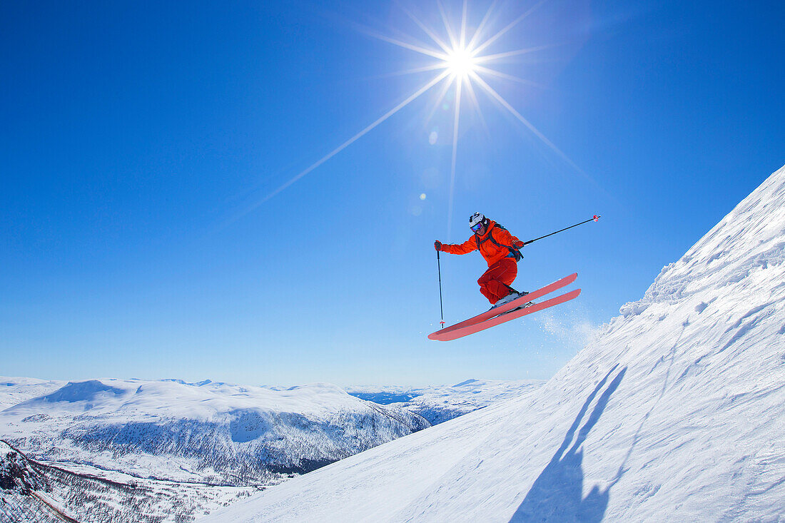 A male freerider in a red suit is jumping from a snow ridge. The sun is shining, the sky is blue. Myrkdalen in the Norwegian region of Flord is a secret powder ski paradise. The resort is close to the Voss area and counts an anual snowfall of up to 10 met