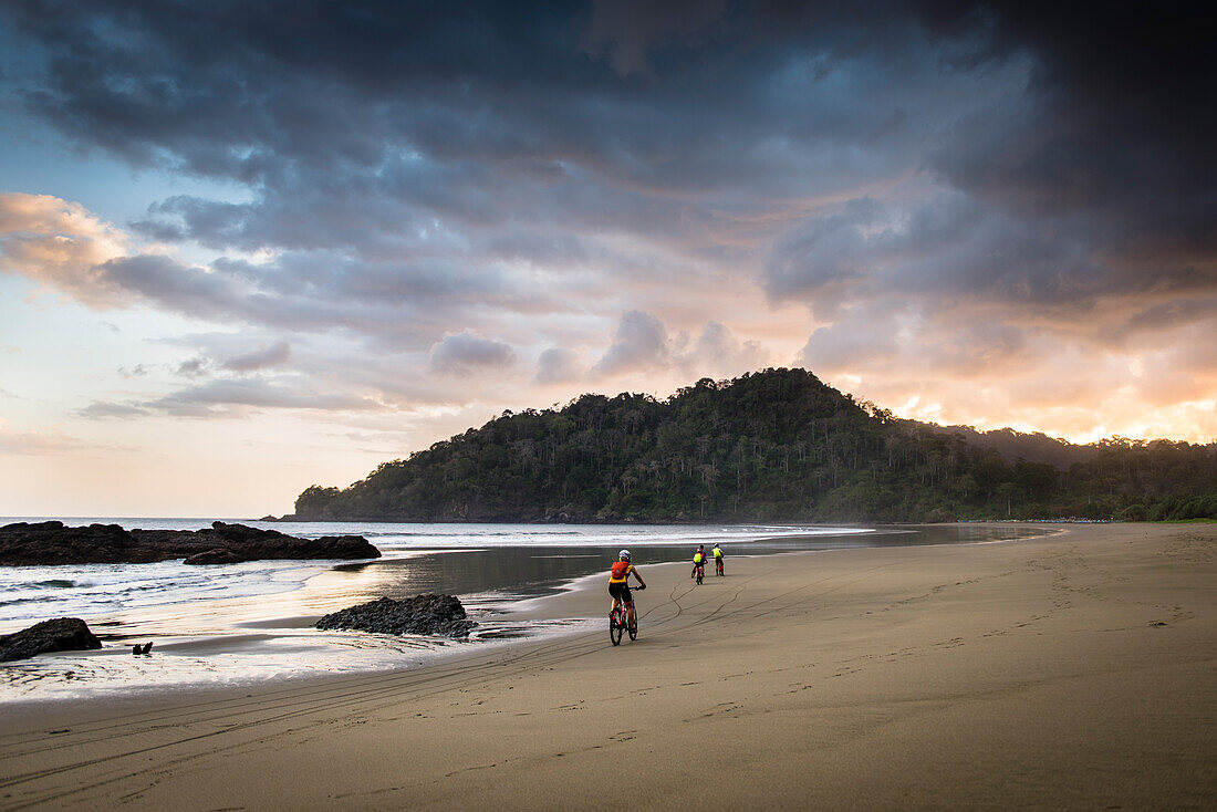 Cyclists riding along a deserted beach towards the sunset, Java, Indonesia