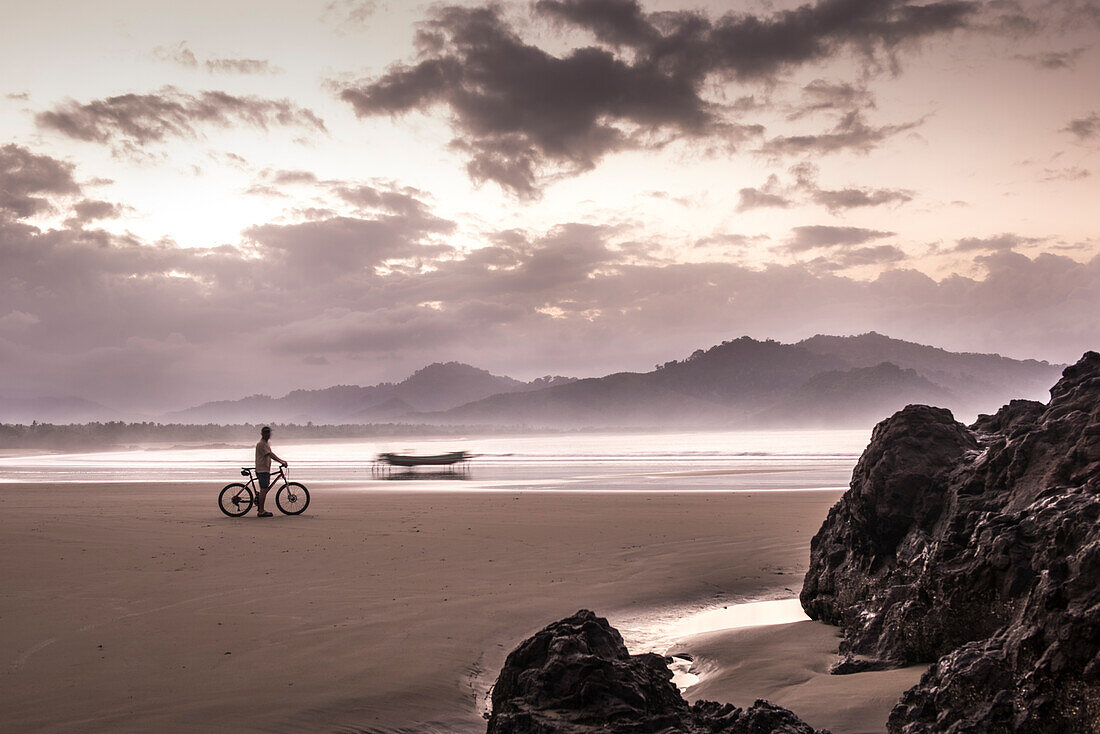 Menschenleerer Traumstrand in einer Bucht mit Radfahrer und Fischerboot während des Sonnenaufganges, java, Indonesien