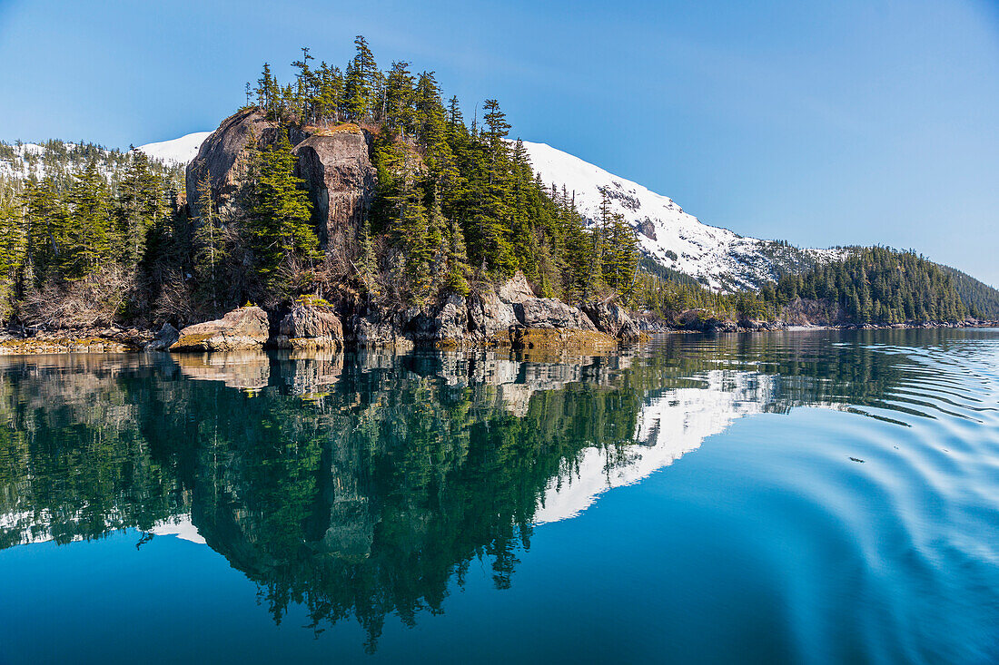 A tall outcropping of rocks and evergreen trees reflects on the waters of Prince William Sound, Whittier, Alaska, United States of America