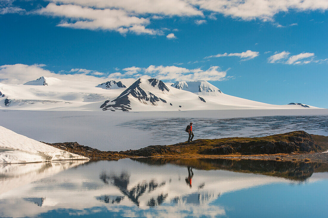 A man hiking by a lake with the Harding Icefield and the Nunatak peaks in the background, Kenai Fjords National Park, Southcentral Alaska