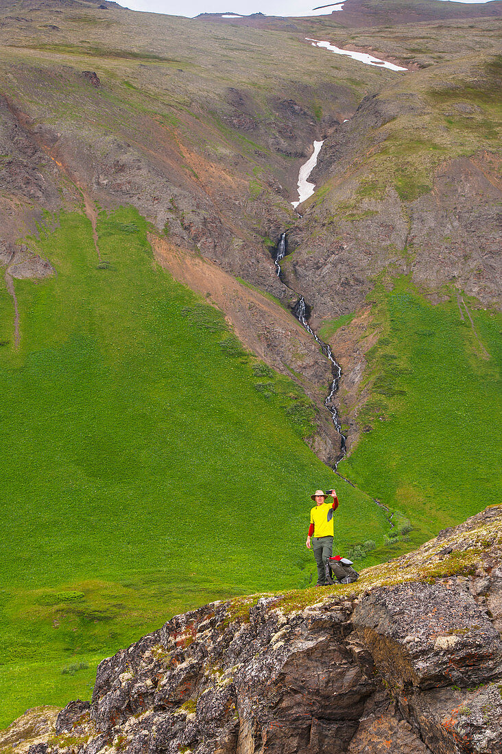A man hiking in Hanging Valley takes a selfie with a waterfall in the background in South Fork near Eagle River, Southcentral Alaska, summer