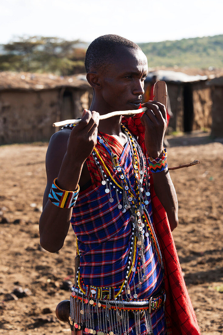 A young Masai man dressed in a traditional costume of blue, black and red checked clothing pretends to play the flute with a stick in the middle of his village, Narok, Kenya