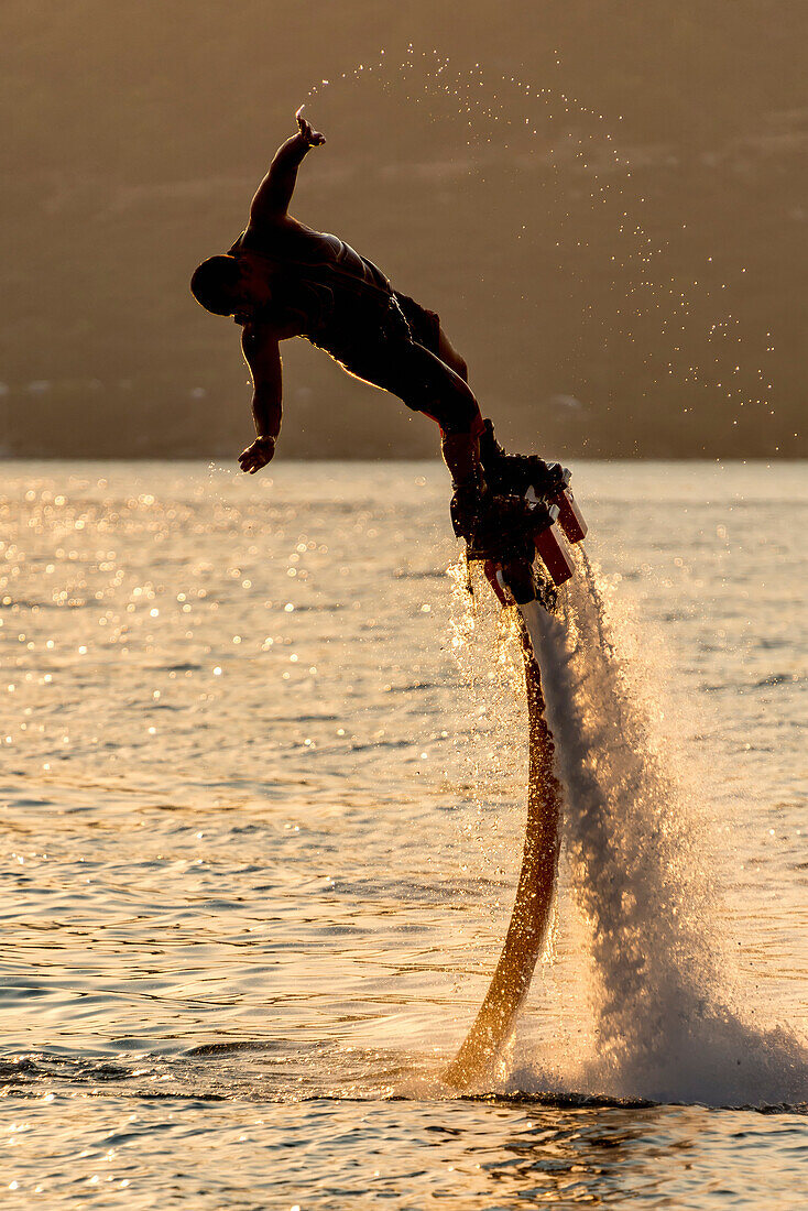 Flyboarder in silhouette leans into dive, trailing water droplets from his hand, Torba, Mugla Province, Turkey