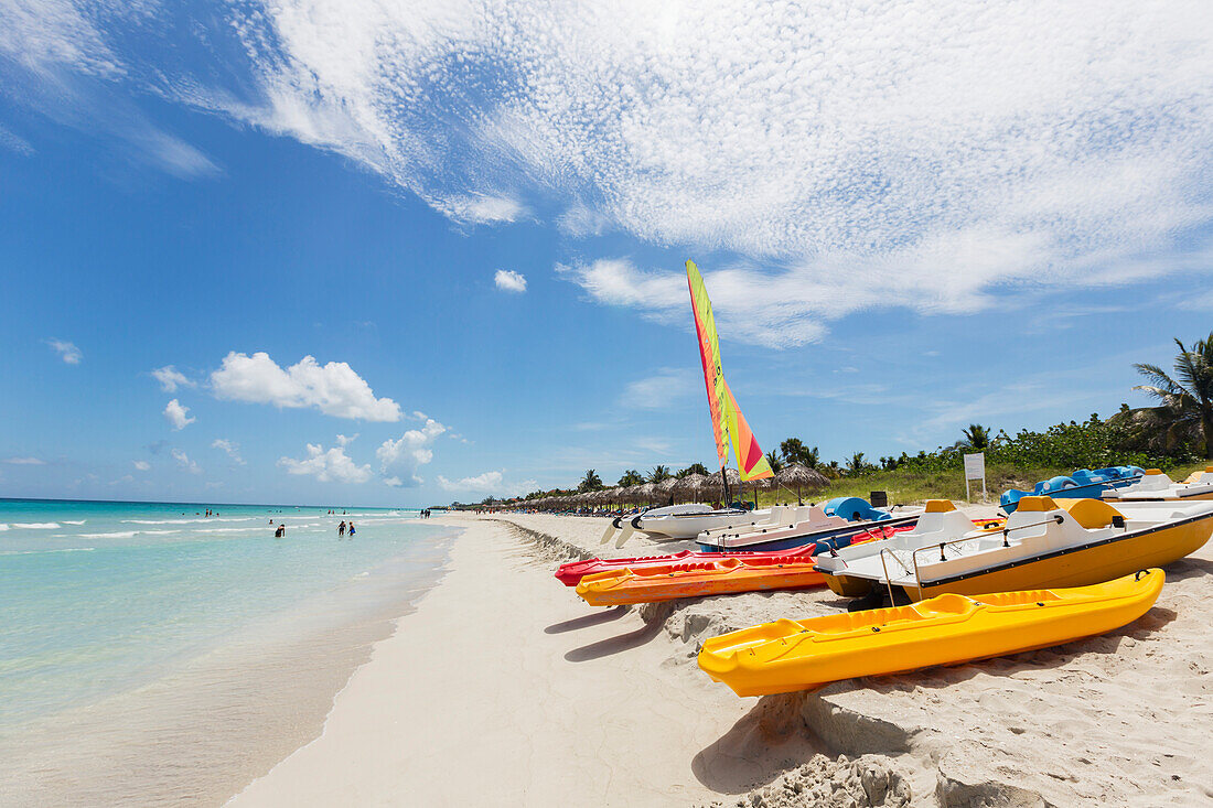 View looking down the white sand beach on a sunny day with kayaks and boats resting in the sand and people in the ocean in the distance, Varadero, Cuba