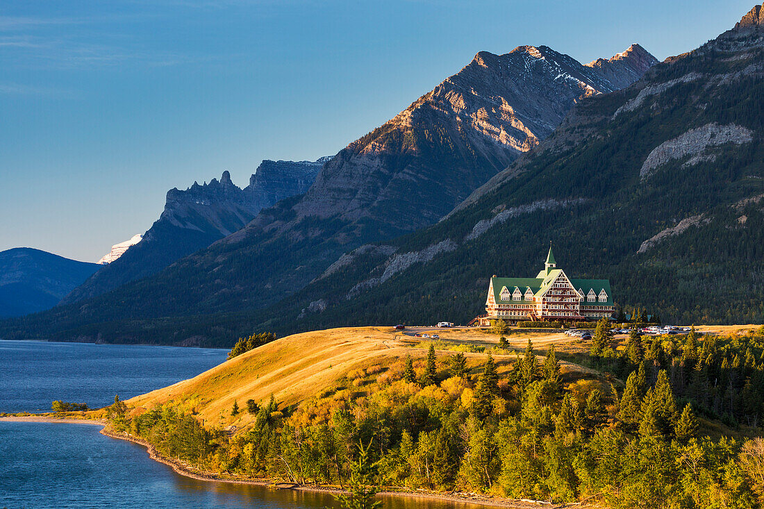 Prince of Wales hotel on hilltop at sunrise overlooking lake with mountain background and blue sky, Waterton, Alberta, Canada