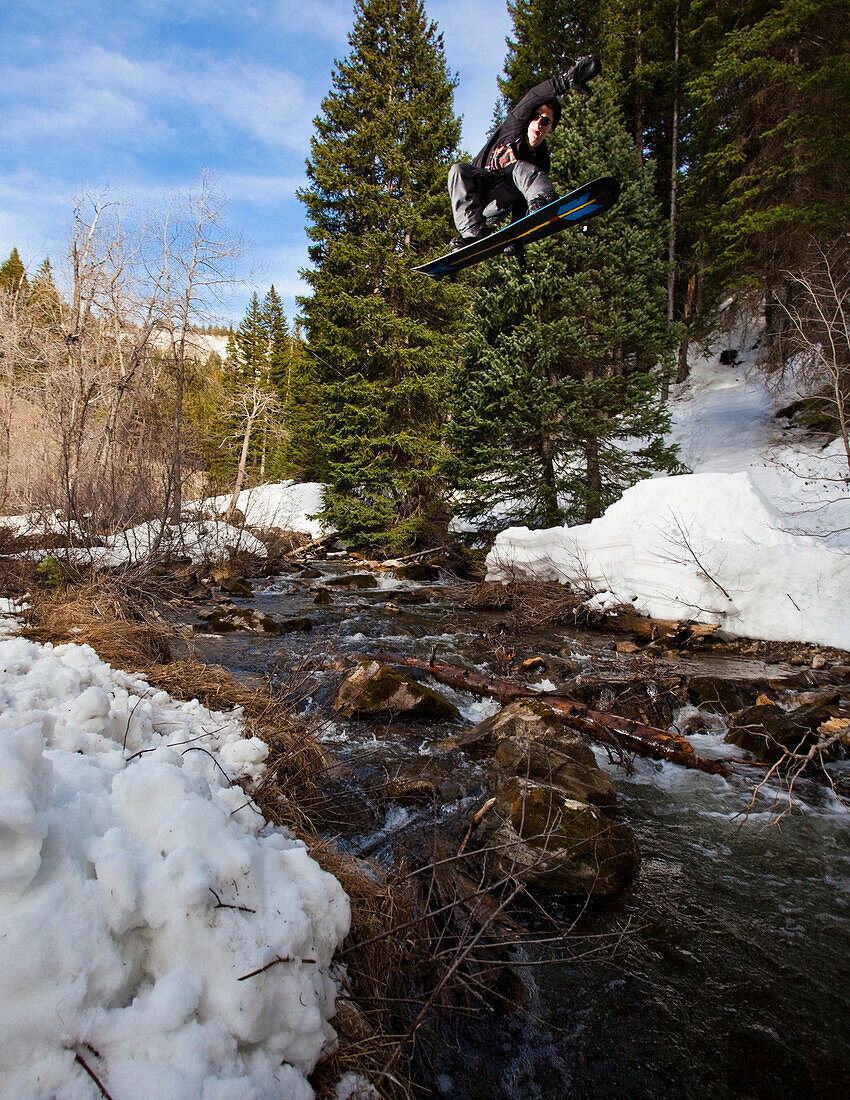 A snowboarder wearing black performs a grab while jumping a creek in Big Cottonwood Canyon.