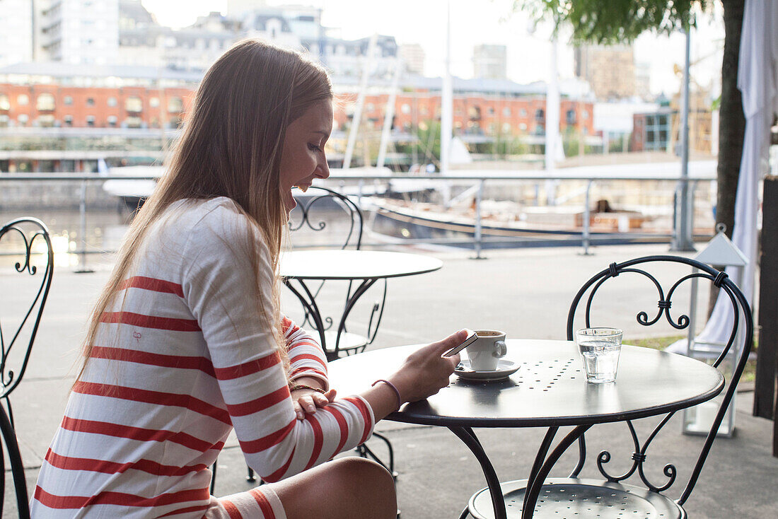 Young woman sitting at sidewalk cafe, laughing at smartphone