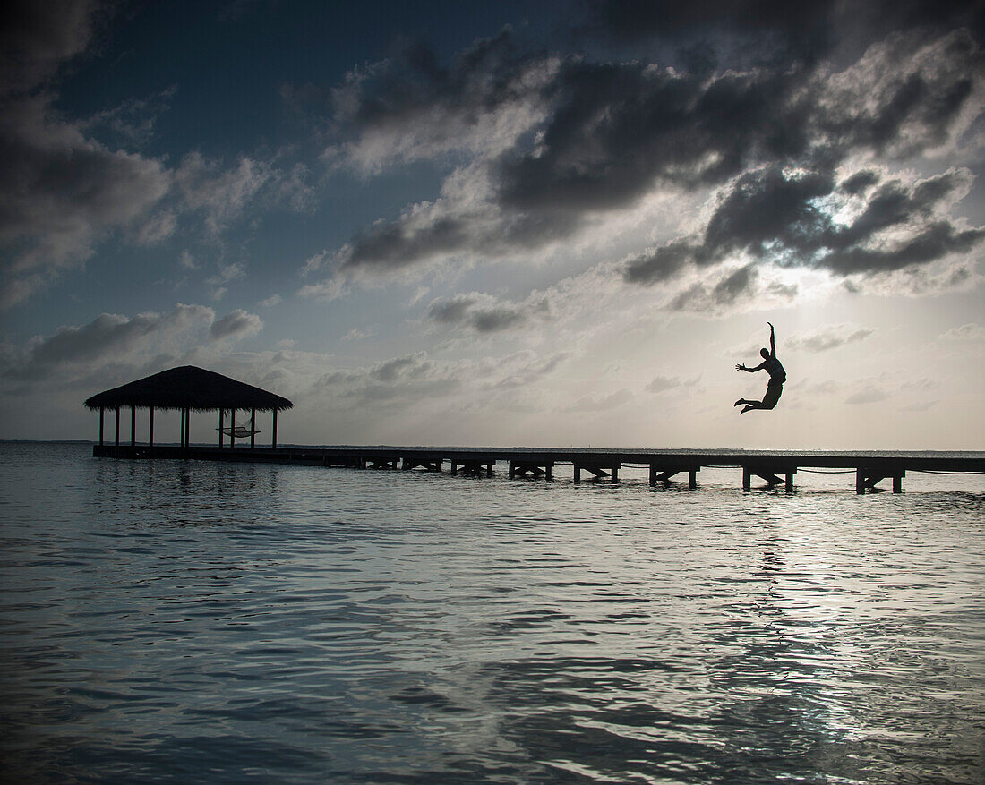 Silhouette person jumping into sea against cloudy sky at dusk