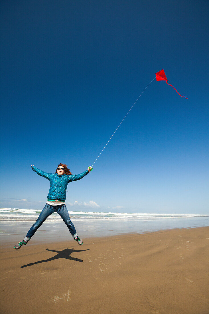 A young woman jumps into the air while flying a red kite in the sky at a beach along the Oregon Coast.