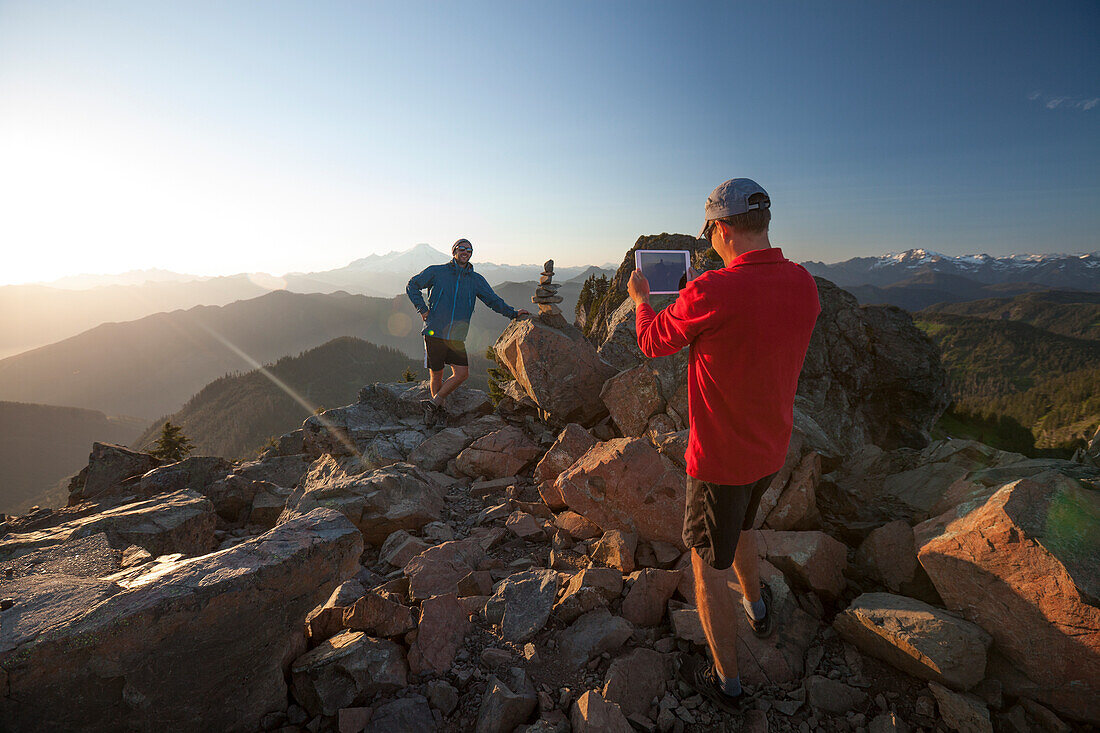 A man uses a tablet to take a picture of his friend on the summit of Sauk Mountain, Washingotn.