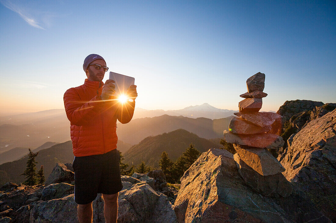 A hiker uses a tablet to take a picture of a cairn on the summit of Sauk Mountain, Washington.