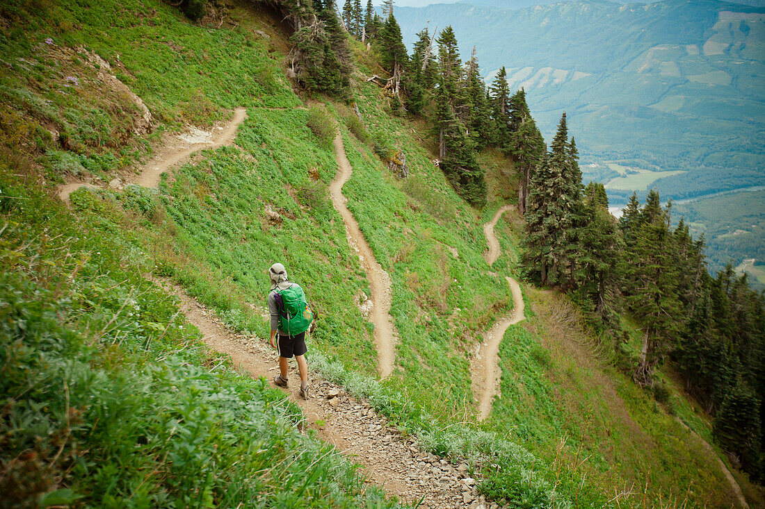 A backpacker hikes along the Sauk Mountain Trail as it switches back and forth through a steep alpine meadow in the North Cascade Mountain Range, Washington.