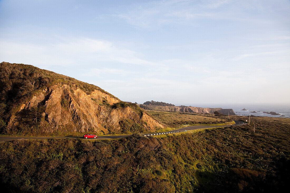 MENDOCINO, CALIFORNIA, USA. A red Prius, a hybrid vehicle, drives on a winding road along the shore of the California coast.