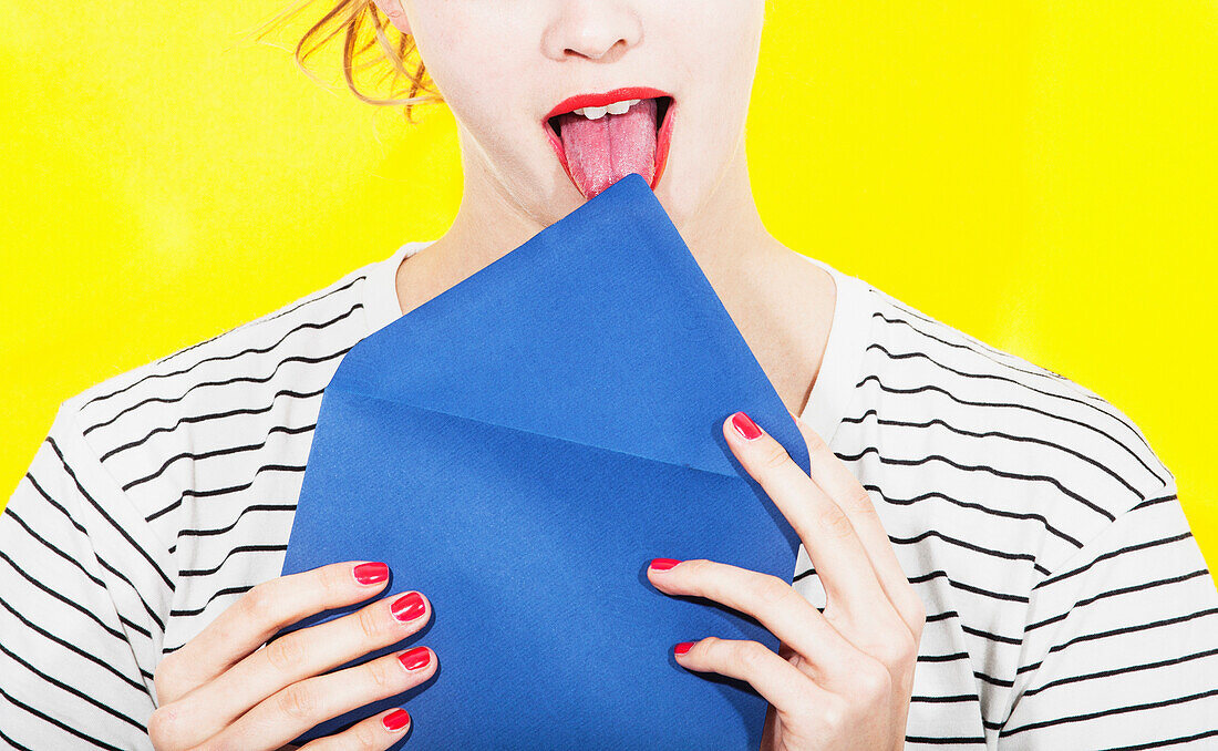 Midsection of woman licking blue envelope against yellow background