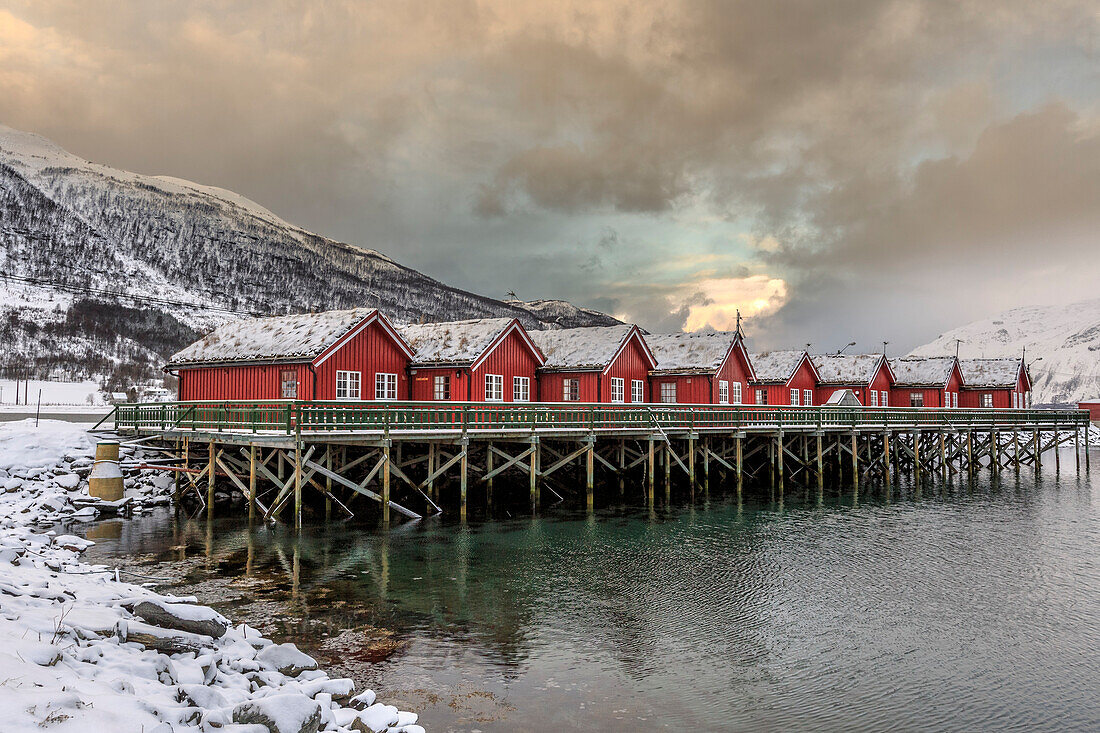 Typical red wooden huts of fishermen in the snowy and icy landscape of Lyngen Alps, Troms, Lapland, Norway, Scandinavia, Europe