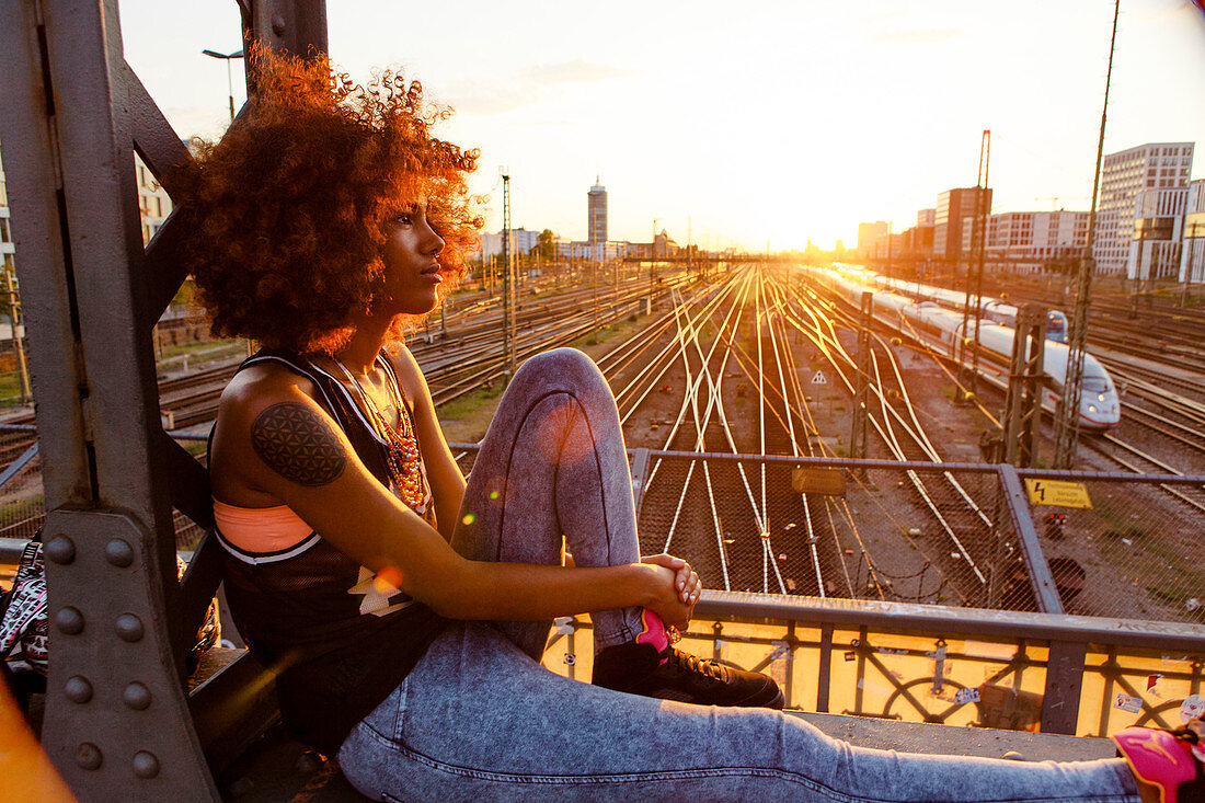 Junge afroamerikanische Frau sitzt im Abendlicht auf Stahlträger in urbaner Szenerie mit Gleisen, Hackerbrücke, München, Bayern, Deutschland