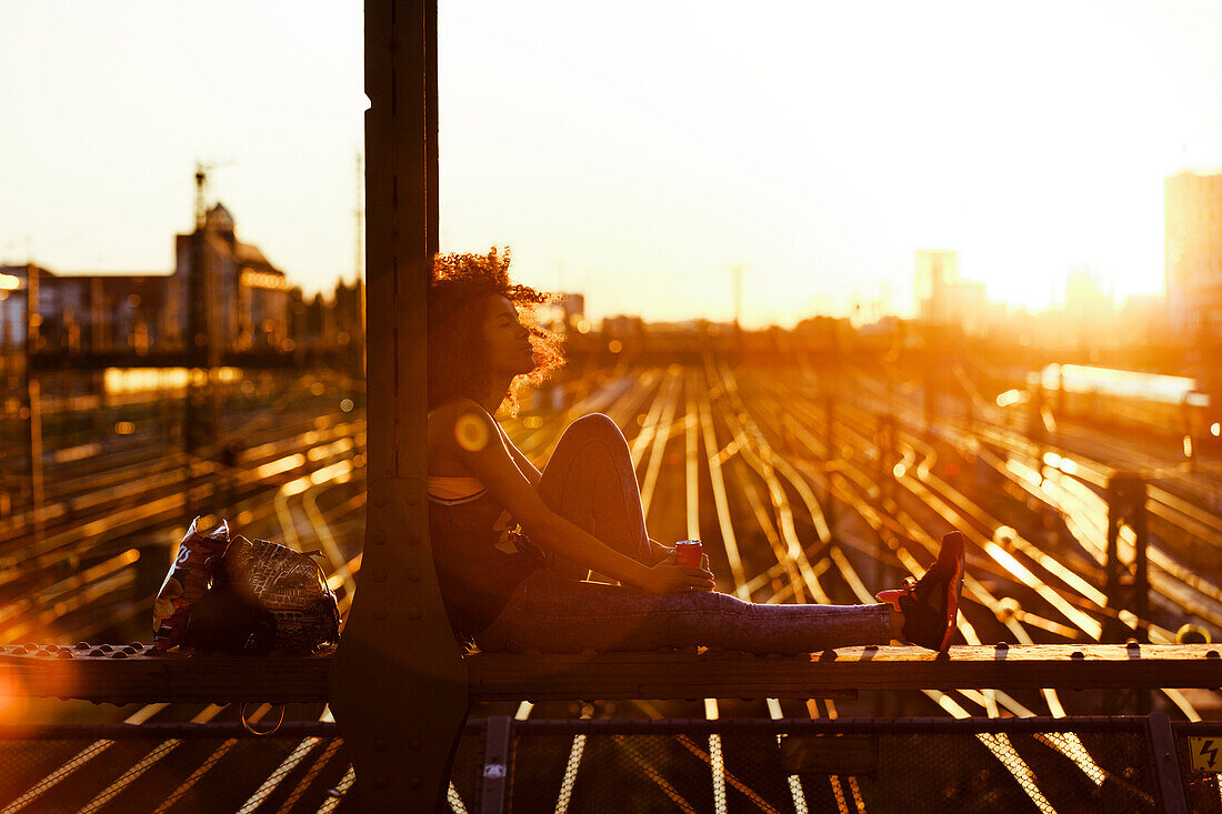 Young afro-american woman in evening light sitting on steel griders in urban scenery with tracks, Hackerbruecke Munich, Bavaria, Germany