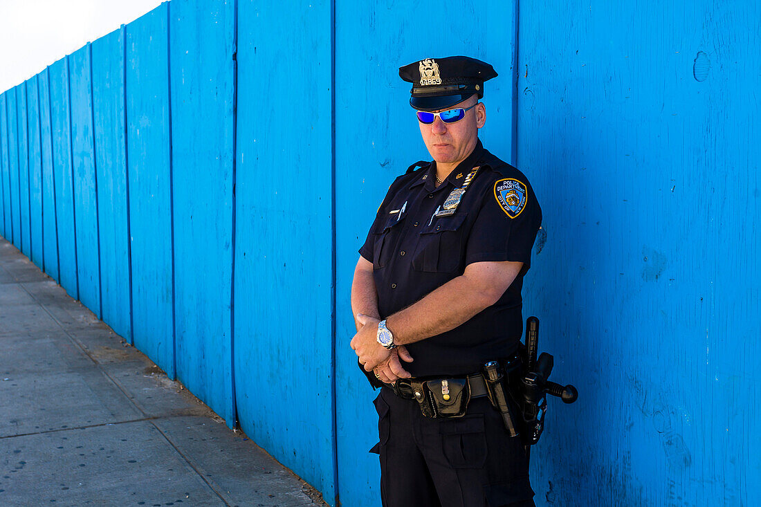 New Yorker Polizist mit voller Ausrüstung und blauer Sonnenbrille vor blauem Zaun schaut ernst in die Kamera, New York City, New York, USA