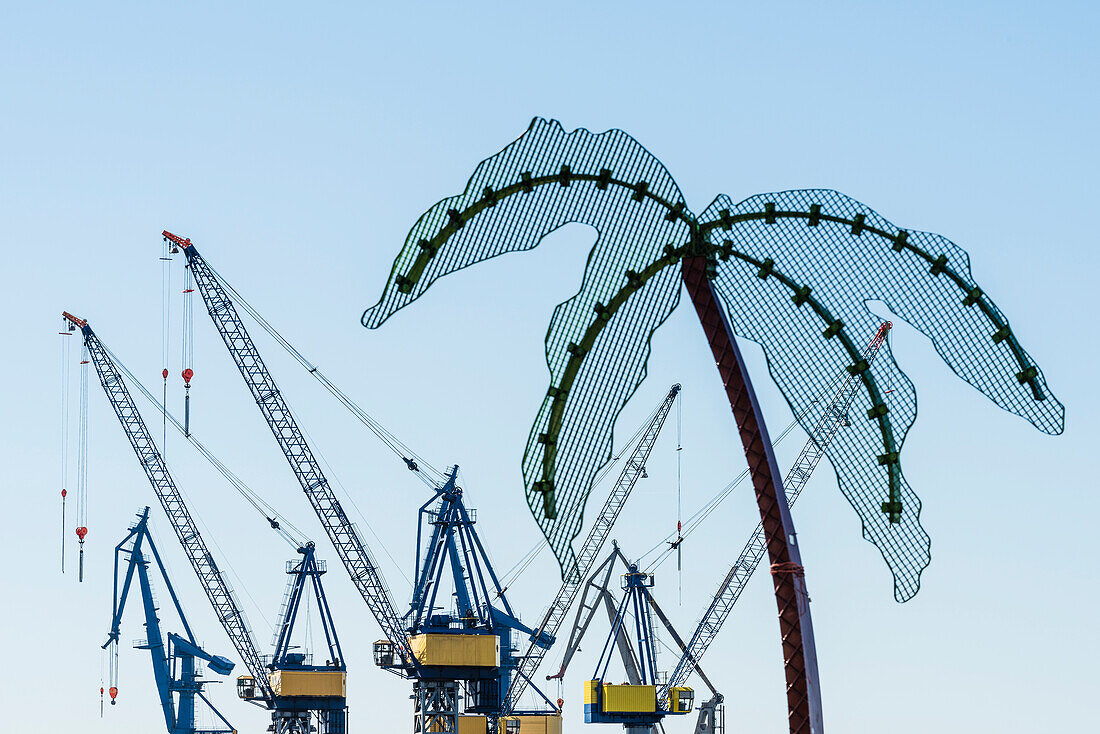 A metal palm tree in the green area Park Fiction on the border Altona Old Town to St. Pauli with look at cranes of the shipyard Blohm and Voss, Hamburg, Germany