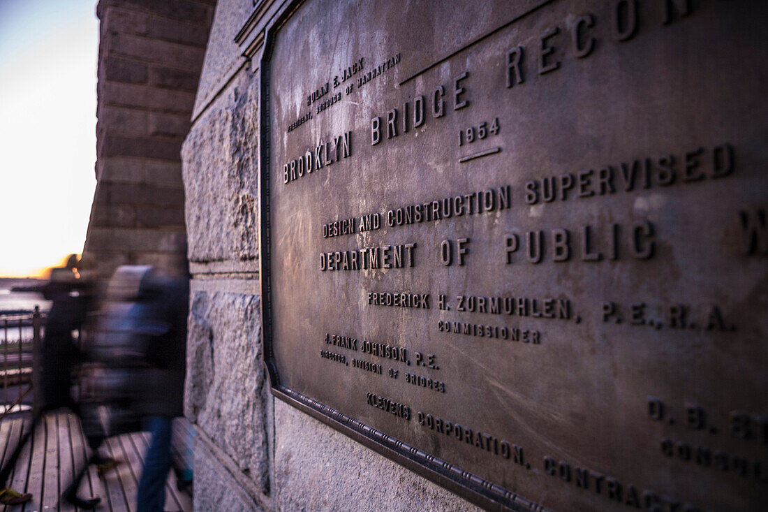 Brooklyn Bridge Reconstruction plaque commemorates the Rebuilding of the bridge in 1954 with Robert Wagner, Mayor. People walk by in background with East River visible.