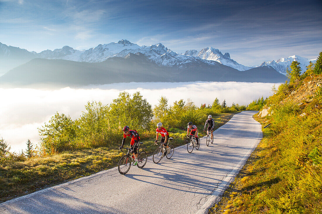 Four road bikers on a winding mountain road on their way to the Emosson reservoir lake in the Alps on the border of France and Switzerland. It is an early morning training session of the local cyclists. The sun is rising, clouds are in the valley, and in 