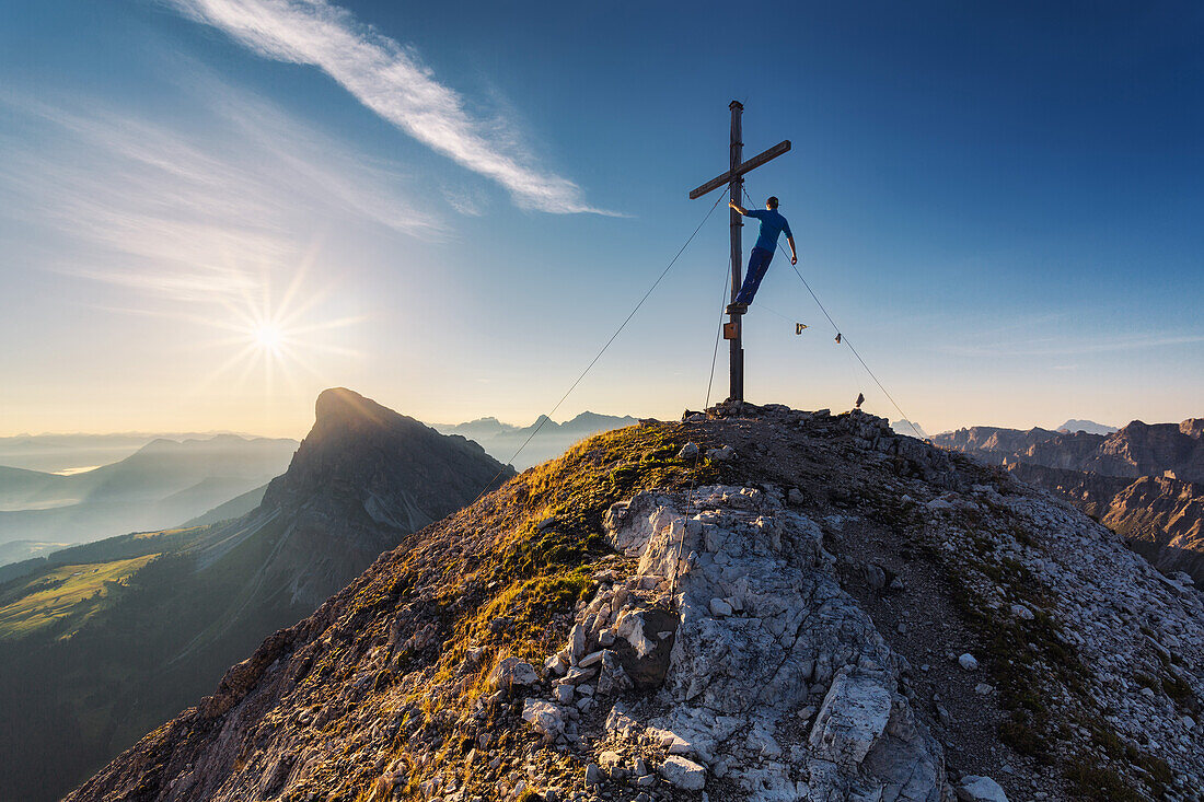 Wanderer klettert auf Gipfelkreuz des Tulln und schaut der Sonne entgegen, Sonnenaufgang in den Dolomiten, Unesco Weltkulturerbe, links im Hintergrund der Peitler Kofel, Italien
