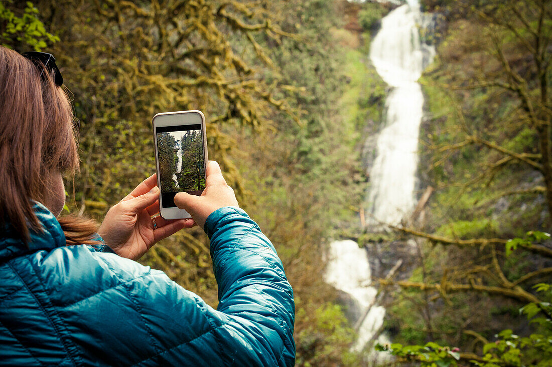 A young woman takes a picture of Munson Creek Falls with her smartphone in Munson Creek Falls State Natural Site