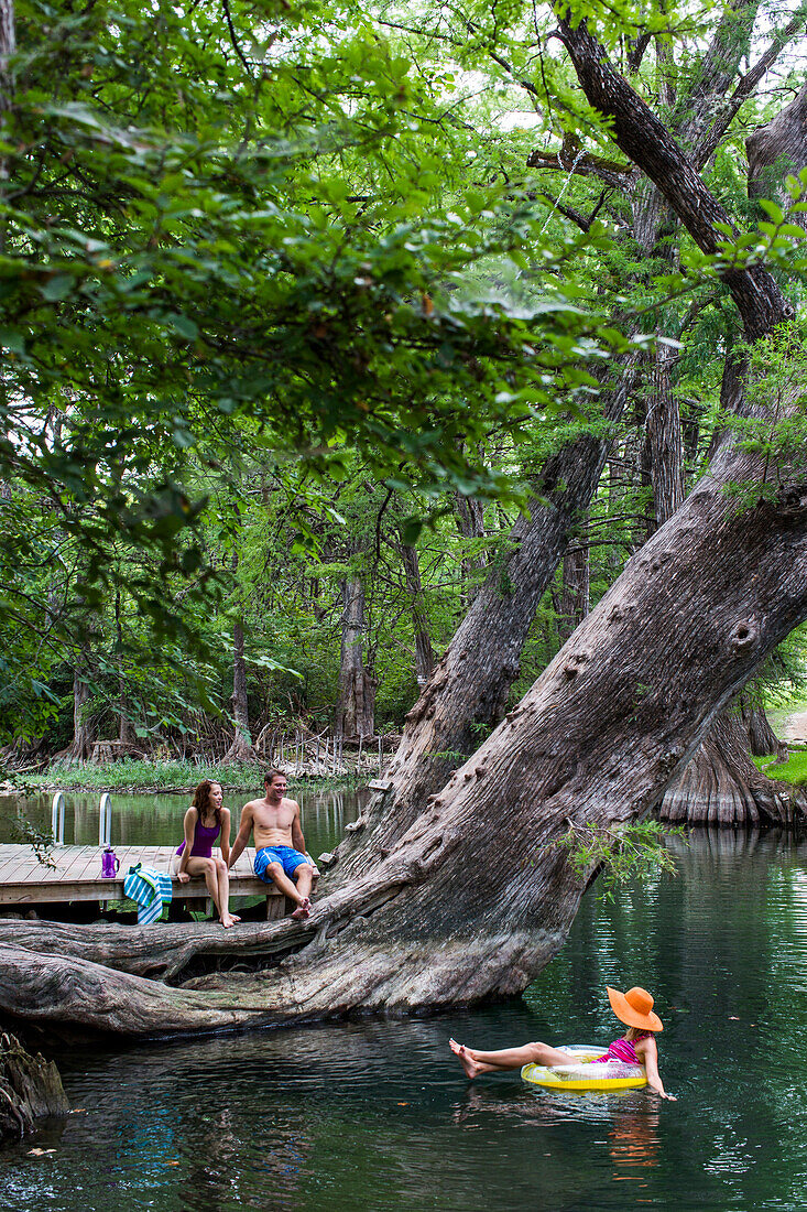 A young woman floats in the water as a man and a woman watch from the base of a tree at the Blue Hole in Wimberley, Texas, a popular destination for tourists and locals on hot summer days. The clear, cool water flows through cypress trees and offers a ref