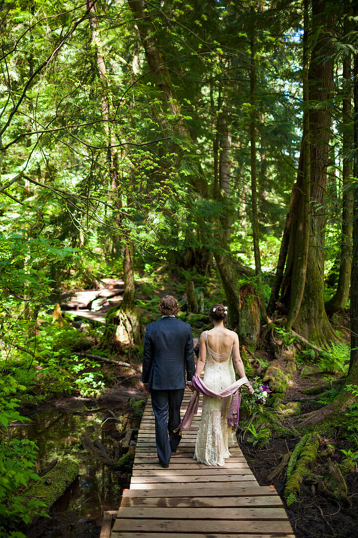 A young married couple hold hands while walking across a boardwalk in the forest.