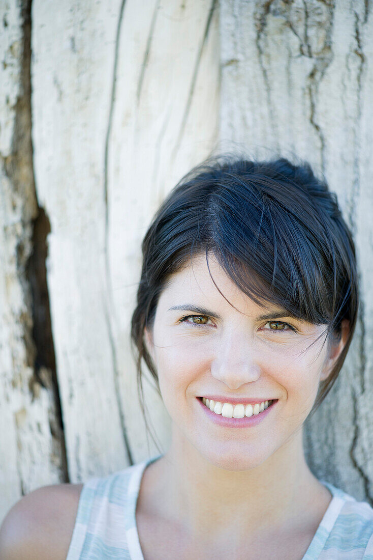 Woman leaning against tree trunk, smiling cheerfully, portrait