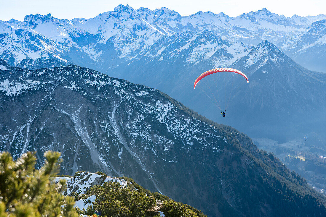 Blick vom Gipfelkreuz des Rubihorns auf verschneiten Wanderweg in Richtung Nebelhorn mit Paragleiter am Himmel, Oberstdorf, Oberallgäu, Deutschland