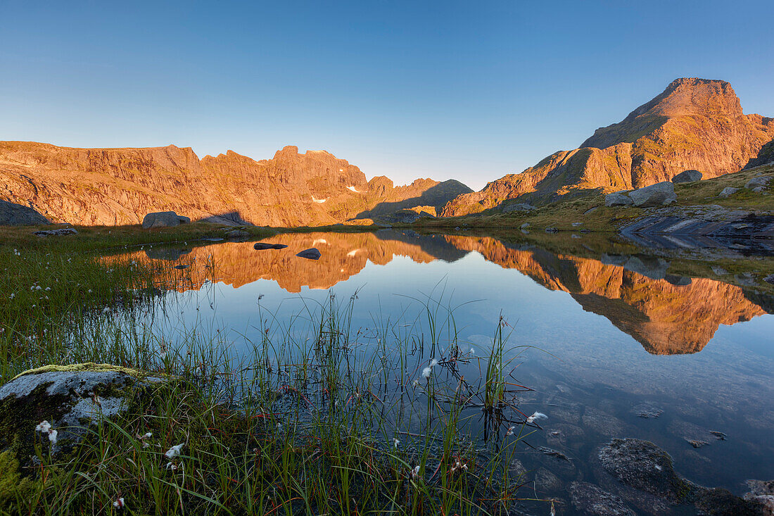 Sonnenaufgang über der vordersten Lofoteninsel Moskenesøy mit den beleuchteten Gipfeln von Hermannsdalstinden (1029 m, rechts) und Ertenhelltinden (940 m, links) und ihrer Spiegelung in einem kleinen Bergsee, Lofoten, Norwegen, Skandinavien