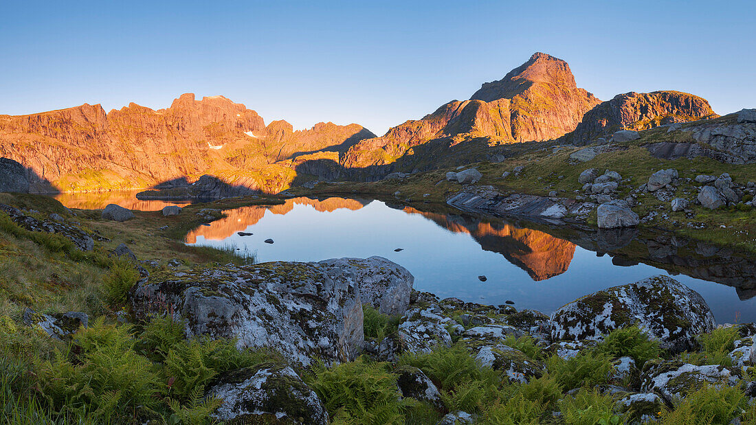 Sunrise over the foremost Lofoten island Moskenesøy with the illuminated peaks of Hermannsdalstinden (1029 m, right) and Ertenhelltinden (940 m, left) and their reflection in a small mountain lake, Lofoten, Norway, Scandinavia