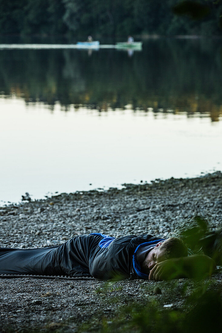 Young man camping at a lake only with a sleeping bag, Freilassing, Bavaria, Germany