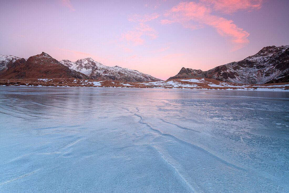 Pink lights of dawn on the snowy peaks around the frozen surface of Andossi Lake, Spluga Valley, Valtellina, Lombardy, Italy, Europe