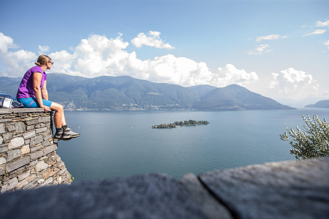 Young female hiker sitting on a wall and looking towards a lake, Ronco, Ticino, Switzerland