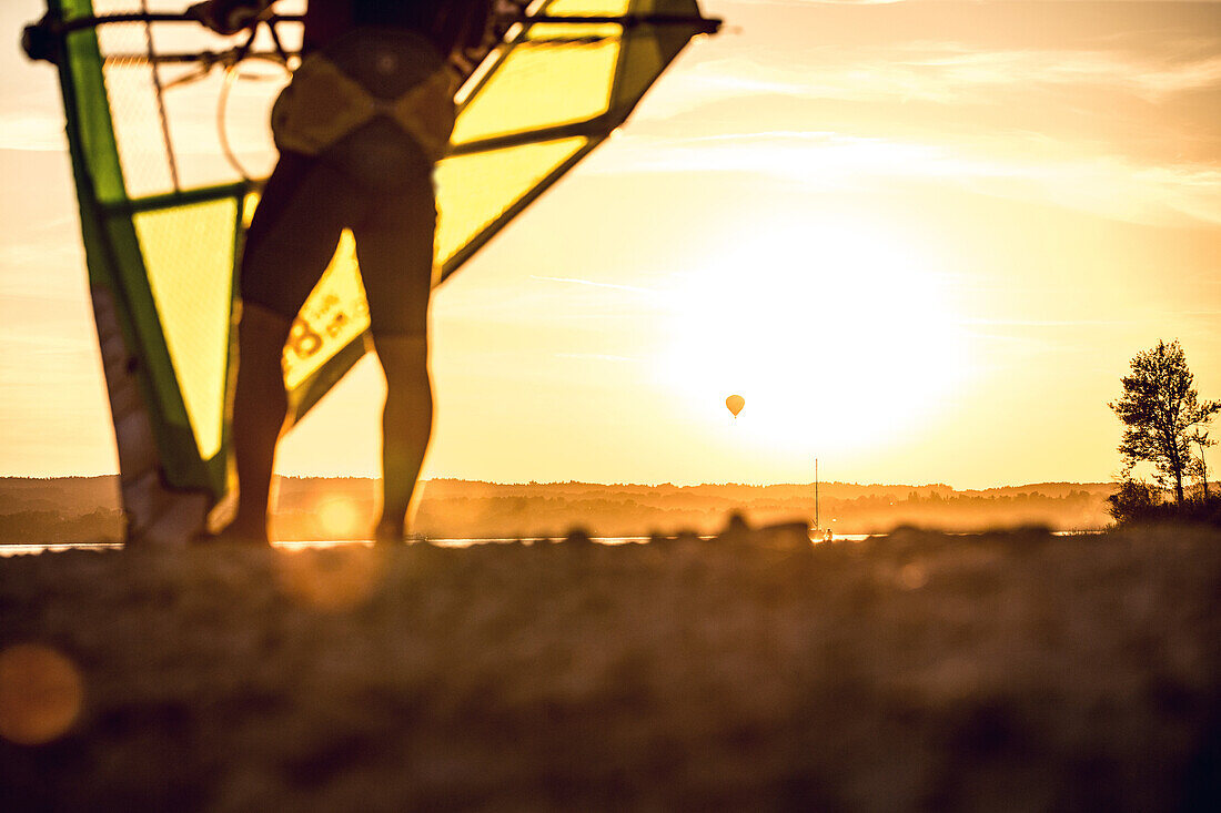 Young male windsurfer checking his sail and looking to a hot-air ballon at a sunny evening, Ammersee, Bavaria, Germany