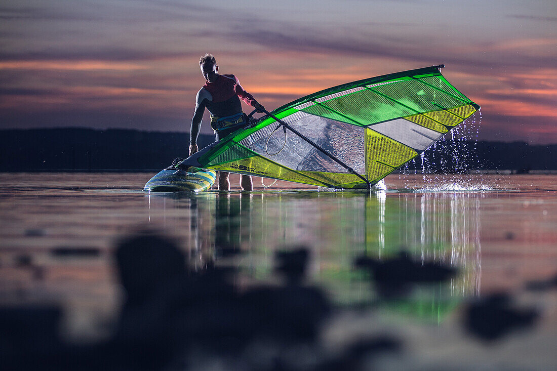Junger Windsurfer traegt sein Segel und Surfbrett aus dem Wasser bei Sonnenuntergang, Ammersee, Bayern, Deutschland