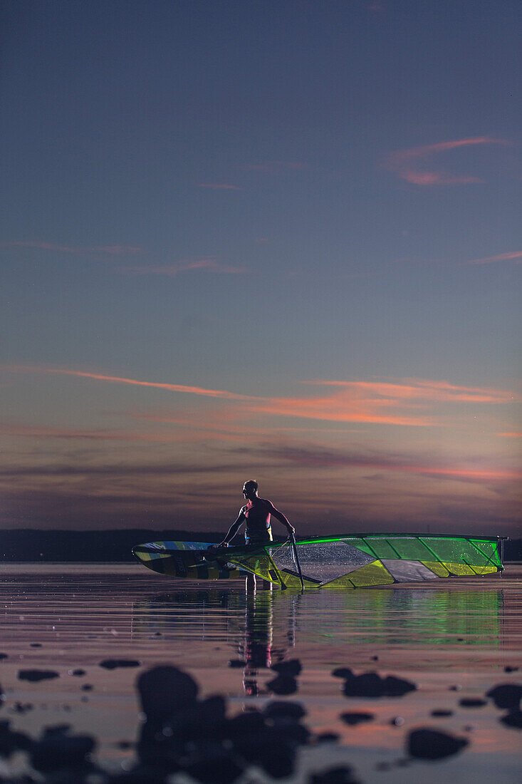 Junger Windsurfer traegt sein Segel und Surfbrett aus dem Wasser bei Sonnenuntergang, Ammersee, Bayern, Deutschland