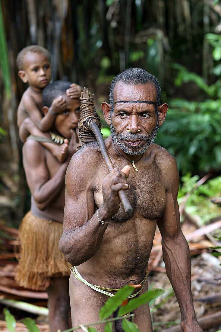 Kombai man standing in the jungle with stone axe his wife with kid in the background, Papua, Indonesia, Southeast Asia.