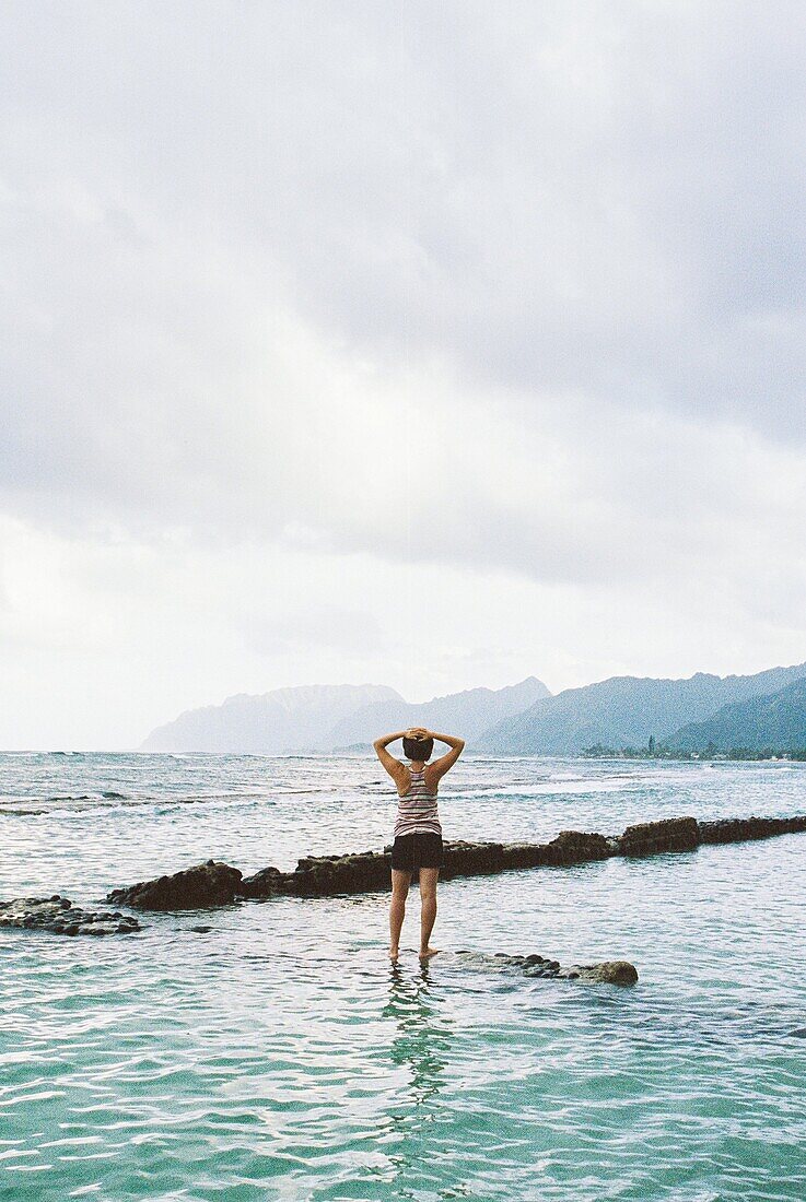 Portrait of a woman standing on rocks with her back to the camera while in the water in Oahu Hawaii.