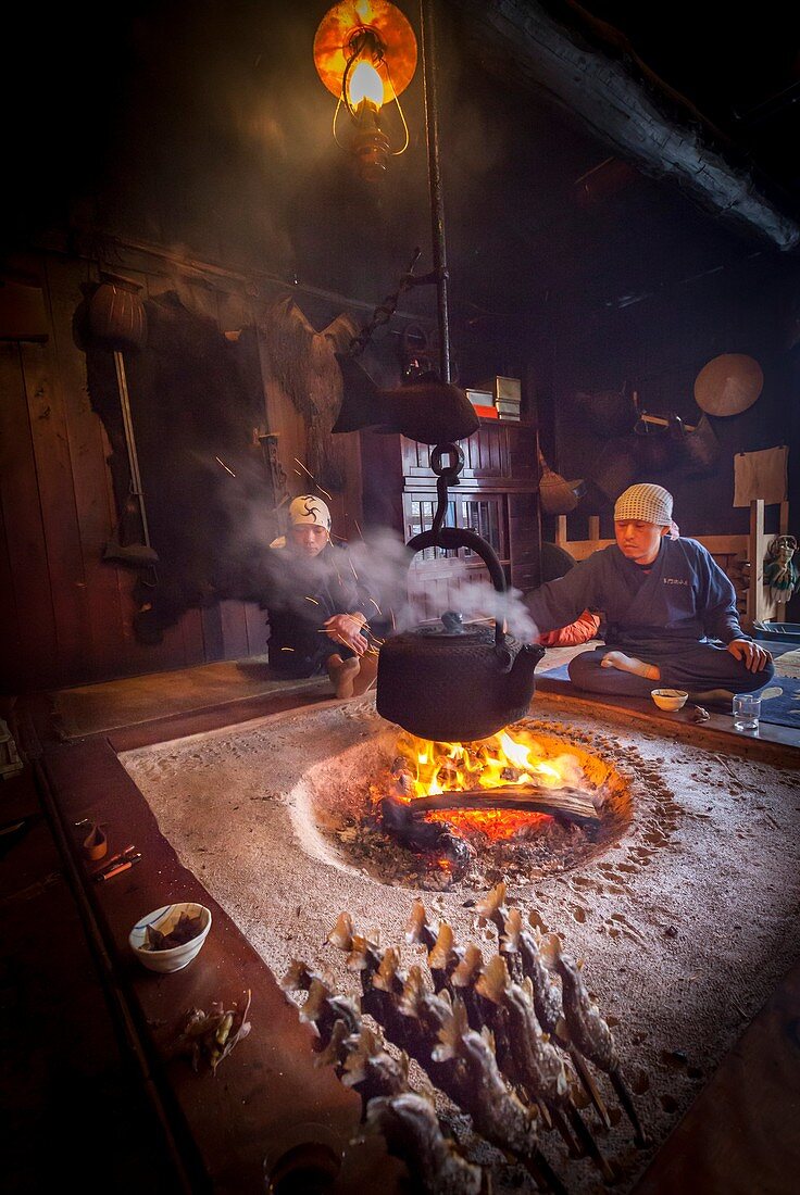Chatting around fire in old mountain lodge, brazed trout on bamboos sticks in foreground, Kamikochi, Northern Japan Alps.