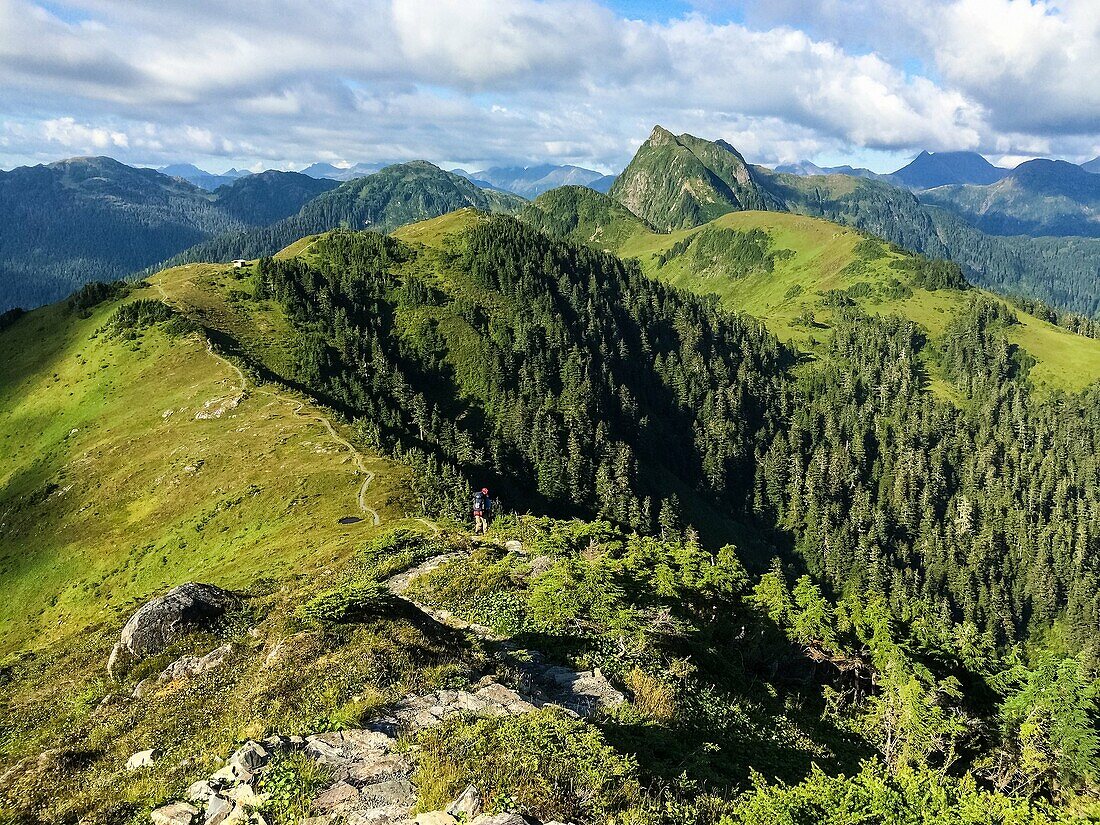 A hiker follows a path on the crest of a bald mountain in Alaska, USA. More of the mountain range can be seen in the distance.