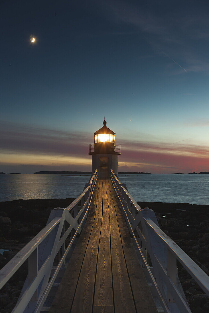 Marshall Point Lighthouse shines its beam into the evening sky to assist mariners entering and leaving Port Clyde Harbor in Port Clyde, Maine. The waxing crescent moon and the planet Venus (just to the right of the lighthouse) follow the sun into the west