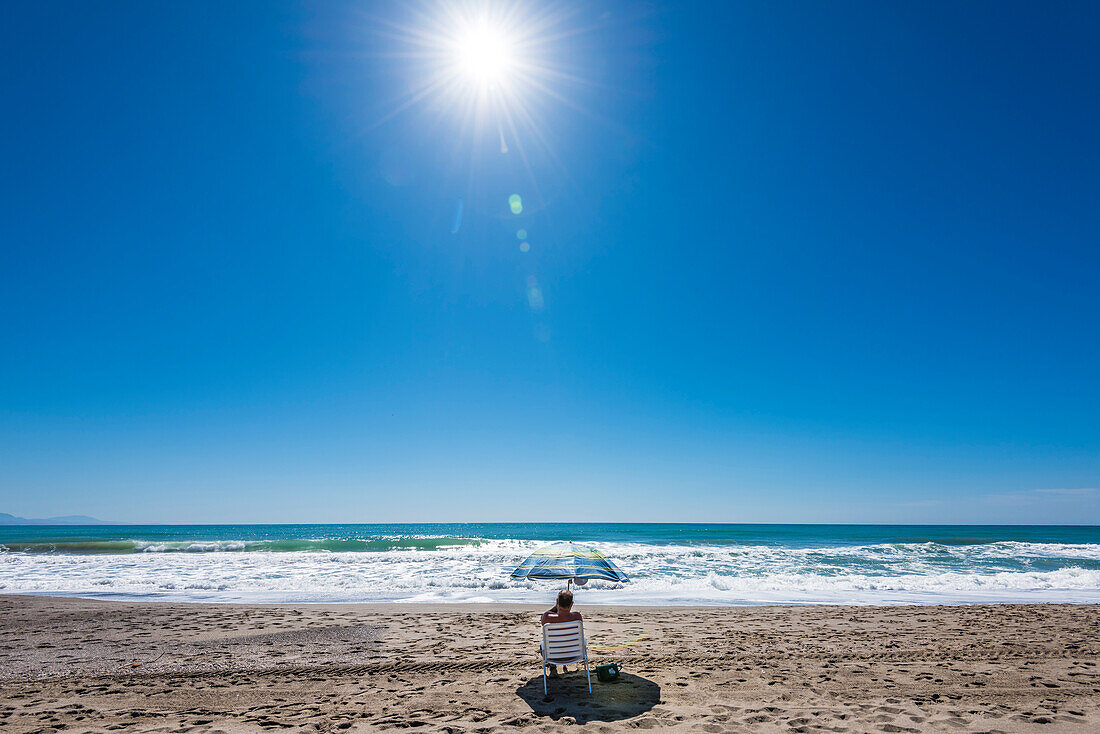 Ein Mann mit Stuhl und Sonnenschirm, bei strahlendem Sonnenschein und blauem Himmel, alleine am Strand, Torremolinos, Andalusien, Spanien