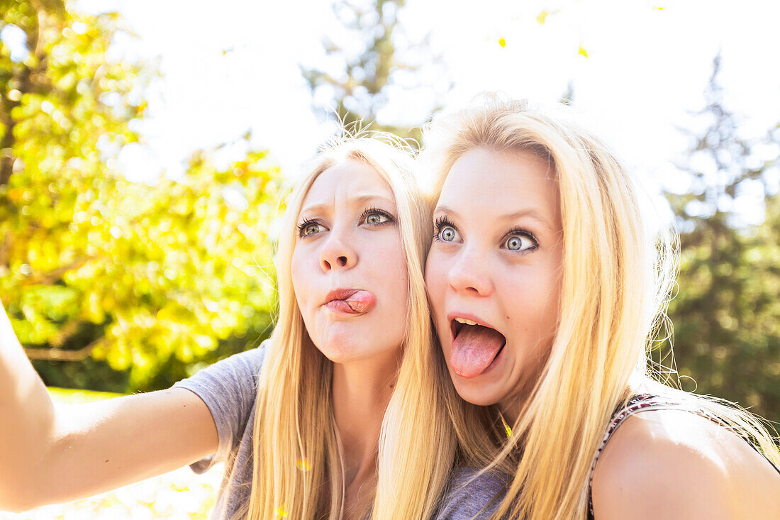 'Two sisters having fun outdoors in a city park in autumn taking selfies of themselves and making funny faces; Edmonton, Alberta, Canada'